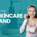 A promotional graphic with a teal background featuring the text "HOW TO START A SKINCARE BRAND" in bold white capital letters. To the right, a smiling woman in a headband and tie-dye shirt applies facial cream from a white jar. The background includes a blurred silhouette of a pump bottle and textured spa towels.